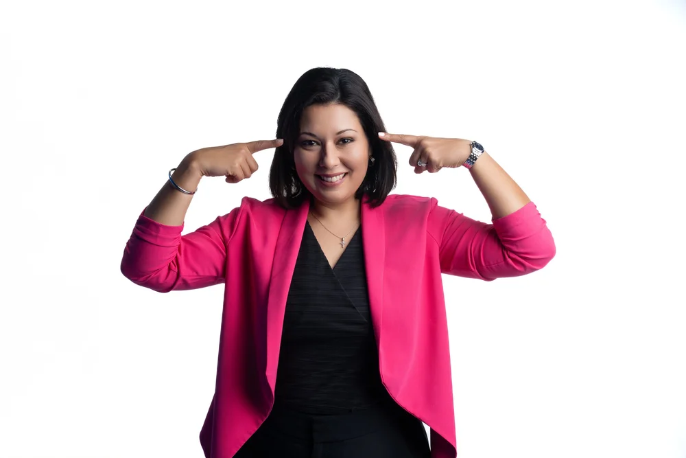 Eva in a pink blazer, smiling and pointing to her head